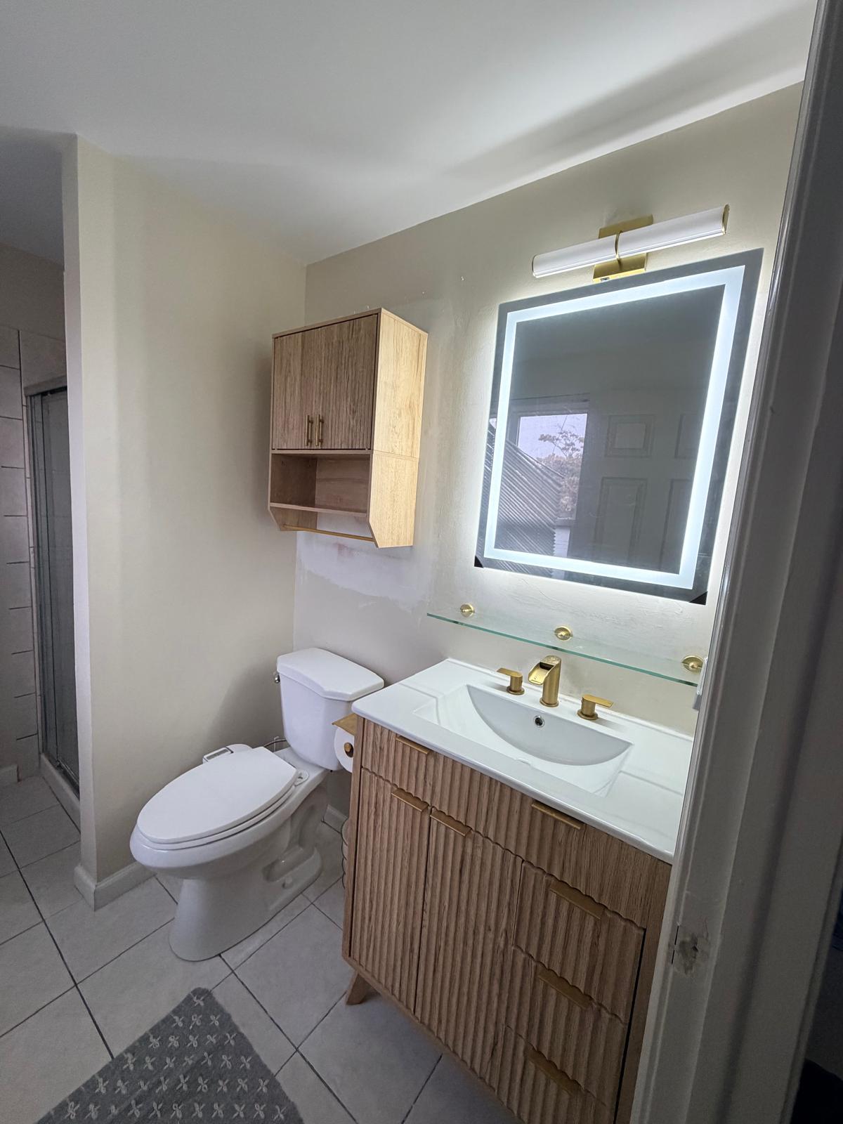 After — bathroom with backlit mirror, wood fluted vanity and cabinet over the toilet and glass shelf