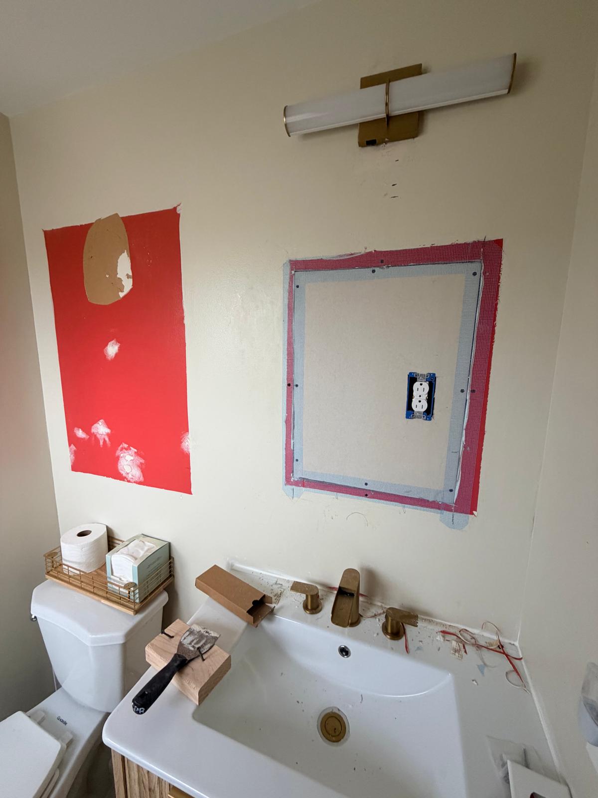 Before — bathroom wall with red drywall patches and repairs, no mirror, tools on the sink